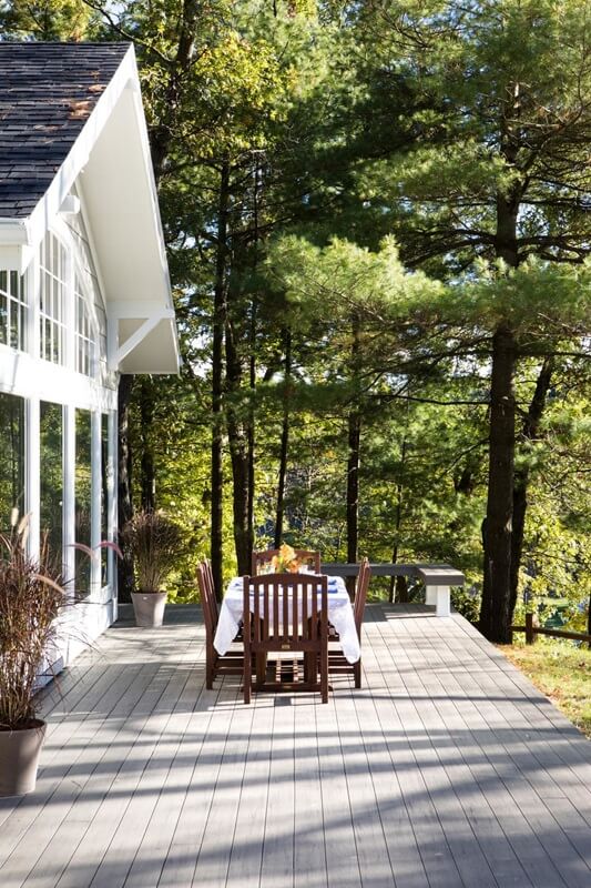 Deck with a table with 4 chair on it. Windows to a house on the left and trees in the background.